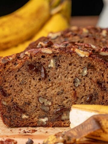 Banana bread with pecans on a piece of parchment paper with bananas in the background.