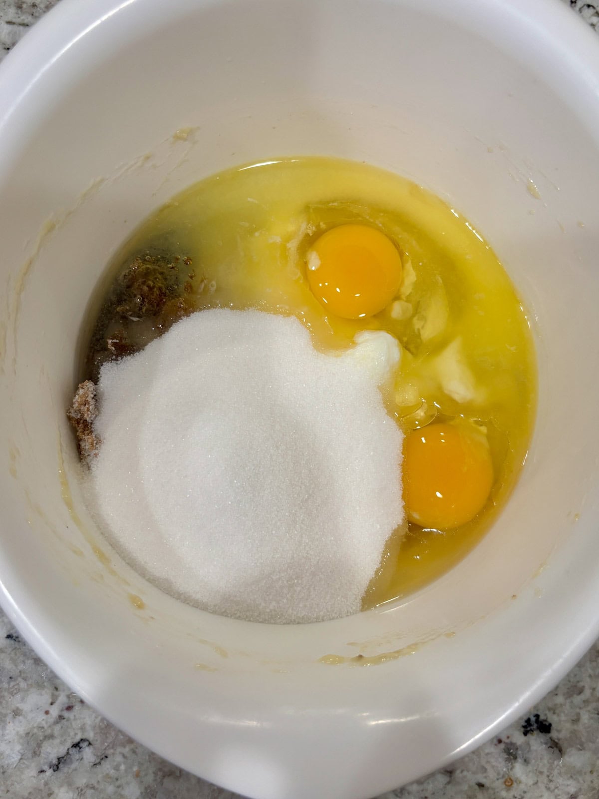 Eggs, banana, sugars, oil, and sour cream in a white bowl to be mixed.