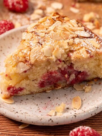 A slice of raspberry and almond cake on a plate with sliced almonds and fresh raspberries around.
